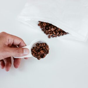 A close-up of a hand holding a measuring spoon filled with coffee beans next to a white bag.
